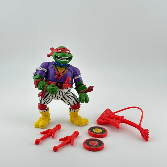 Anthropomorphized turtle wearing zebra print pants, purple shirt, red mask, & yellow boots. Red musical accessories.