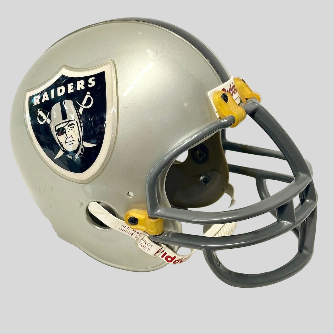 A silver Oakland Raiders football helmet with a dark gray faceguard and yellow plastic fasteners, featuring the team's logo on the side.