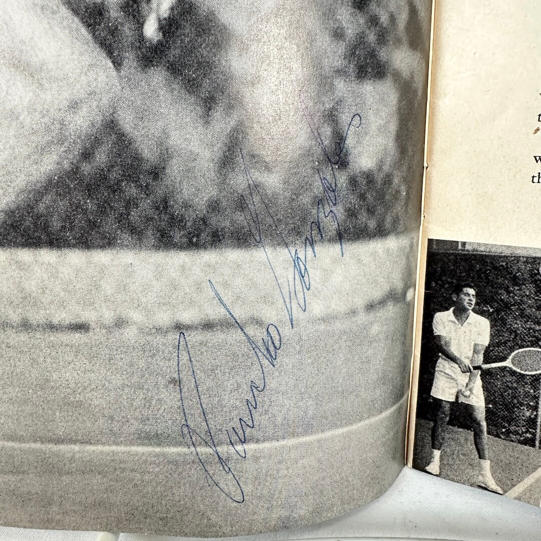 Close-up of a signature on a black and white photo of a person playing tennis in the background.