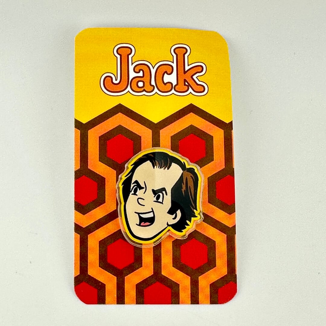 A collectible pin of the character Jack from the movie 'Psycho' on a yellow and orange geometric card backing.