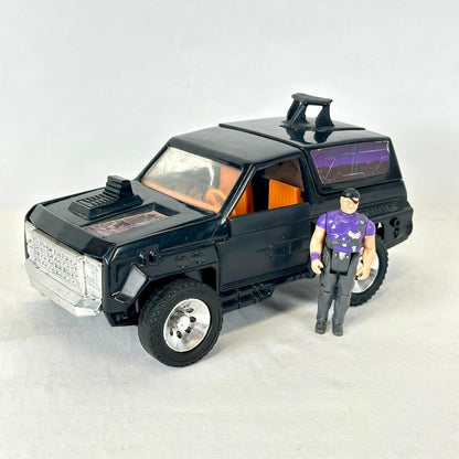 A vintage black toy SUV with orange interior windows and a small action figure standing beside it. 