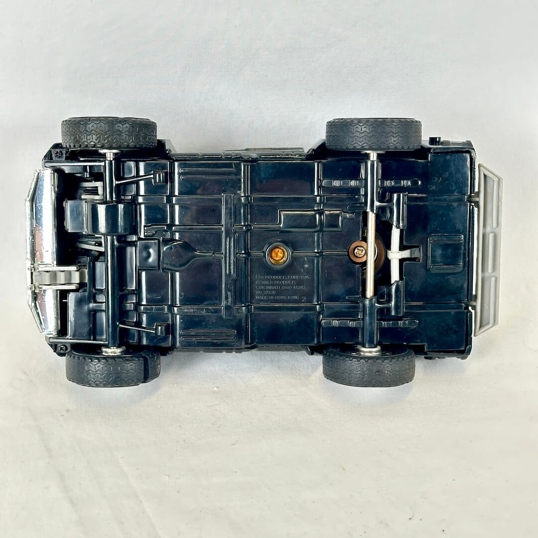A vintage black toy car shown from a top-down view, lying upside down on a light gray fabric background.