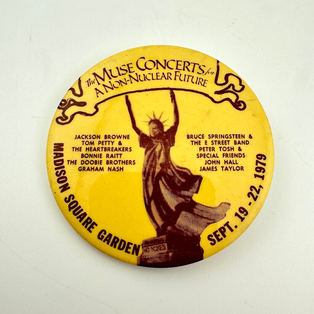 A round yellow vintage concert pinback button featuring a dark brown illustration of the Statue of Liberty and event details text.