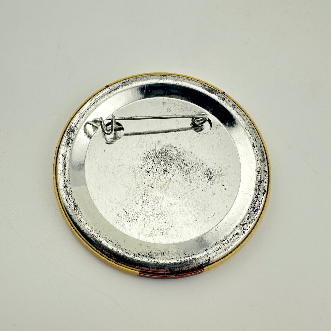 Backside of round pinback badge with yellow rim and silver metal back with bar attachment. 