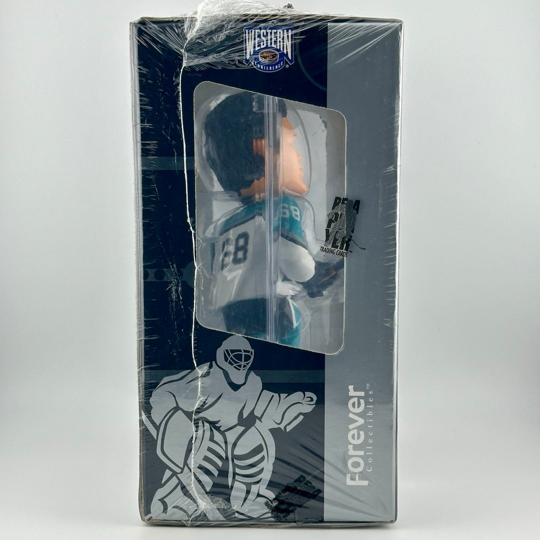 Hockey player wearing teal & white uniform holding stick still in box wrapped in plastic.