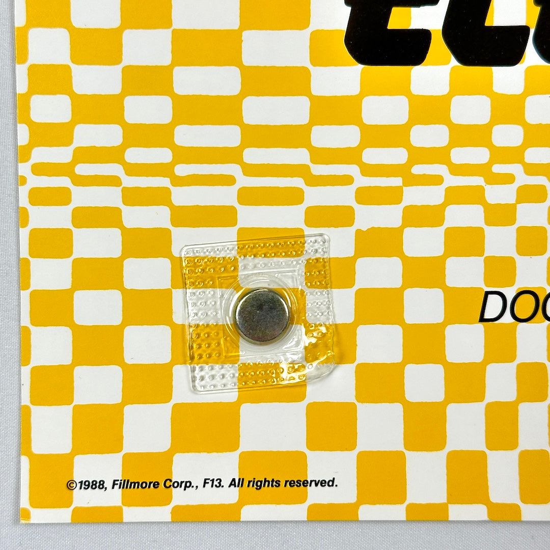 Yellow and white checkered product packaging with a clear square featuring a circular magnet, on a gray background.