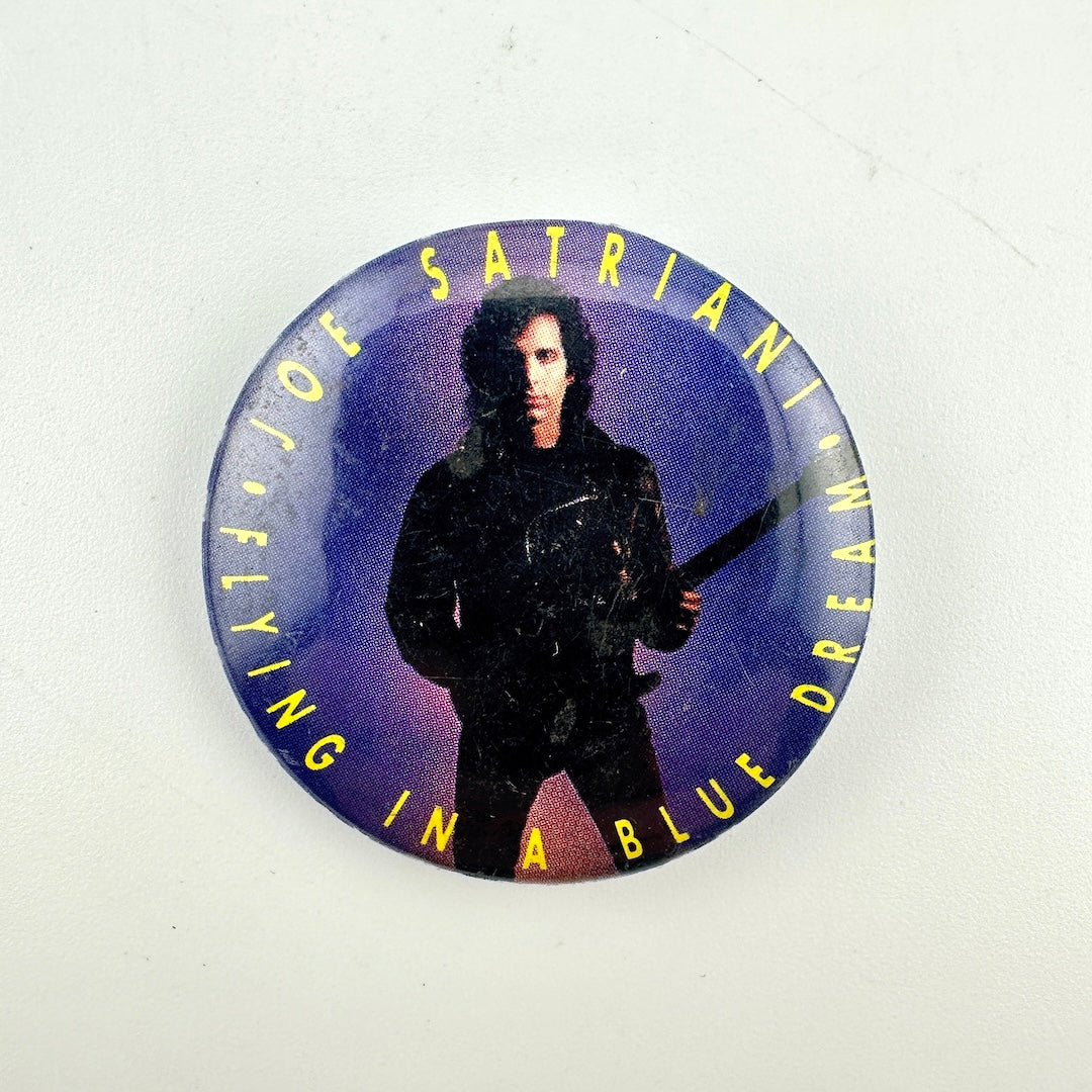 A round button pin featuring a photo of Joe Satriani holding a guitar, with the text in yellow around outside. 