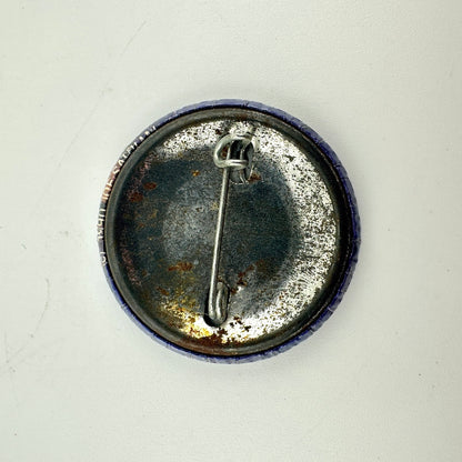 The back of a round pinback button badge, showing a tarnished silver metal safety pin clasp.
