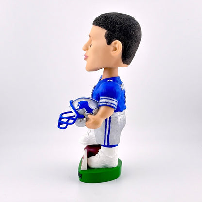 Side view of football player bobblehead in blue jersey holding helmet against white background. 