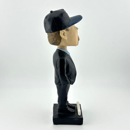 Bobblehead figure of a person wearing a dark suit and cap on a white background