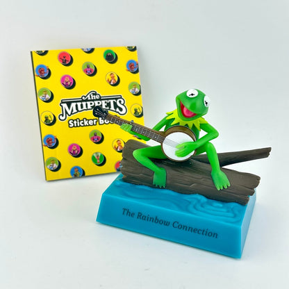 Green frog figurine playing a banjo with a 'The Muppets' sticker book in the background.