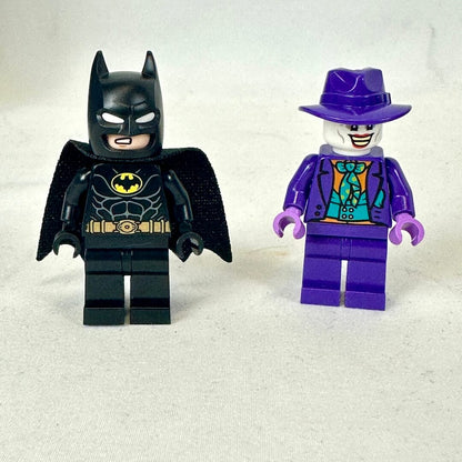 Two LEGO minifigures, one resembling Batman and the other resembling The Joker, on a light gray background.
