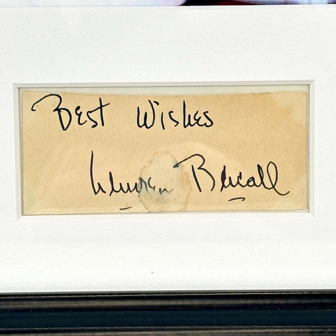 A framed handwritten card with a beige background and black ink writing that says 'Best Wishes, Lauren Bacall"