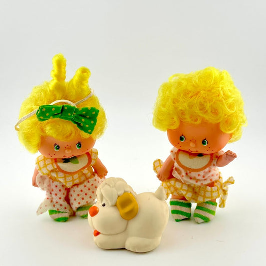 Two vintage dolls with yellow hair and a white toy dog on a light gray background