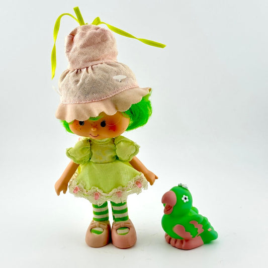 Toy doll in green dress and pink hat with green bird by side against white background. 