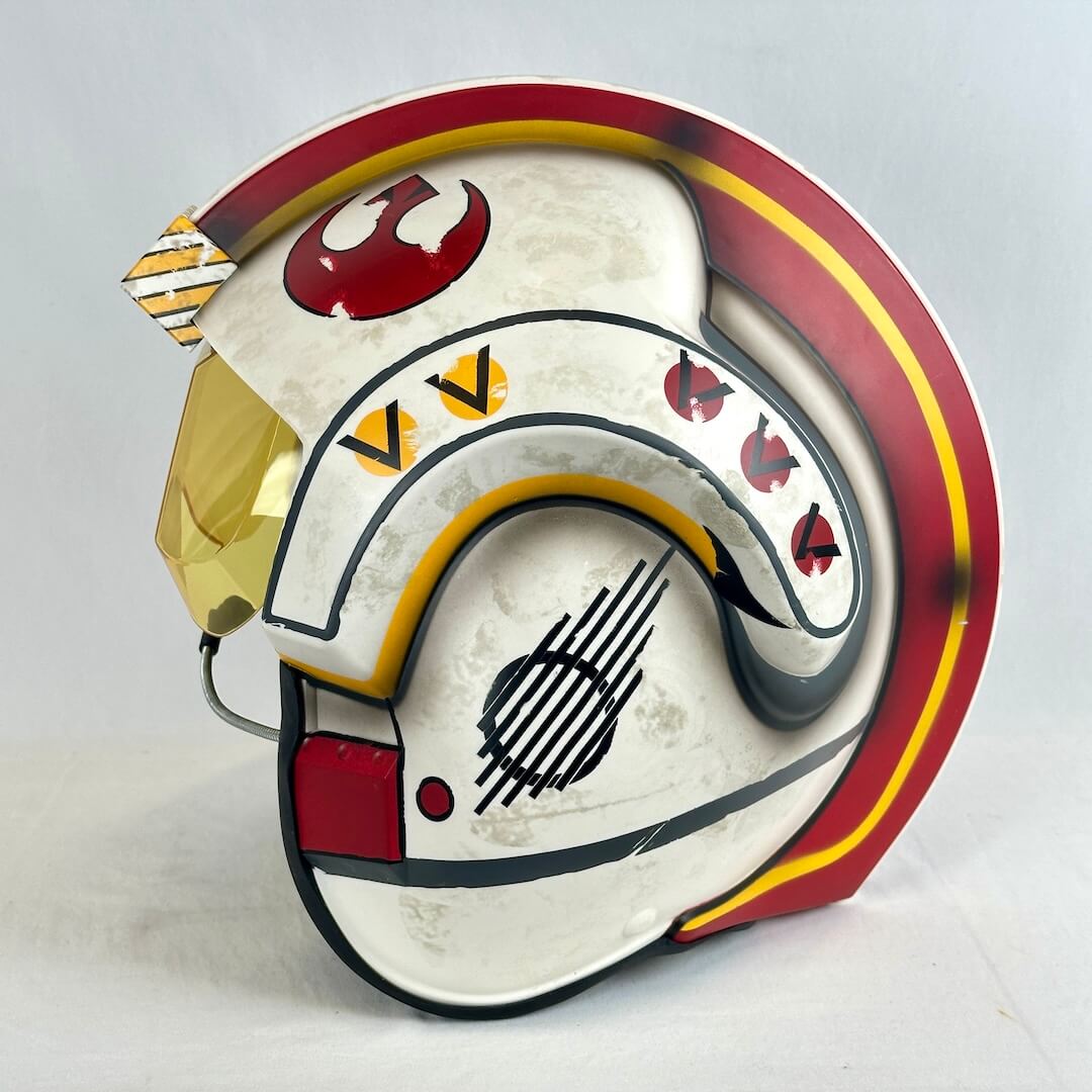 A replica helmet inspired by Star Wars' X-wing, featuring a cream-colored base with red, yellow, black, and gold details.