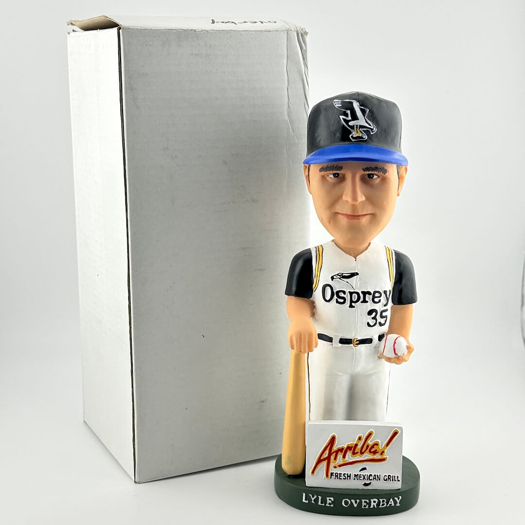 Baseball bobblehead figure with 'Osprey' uniform and 'Arriba' sign in front of a gray box.