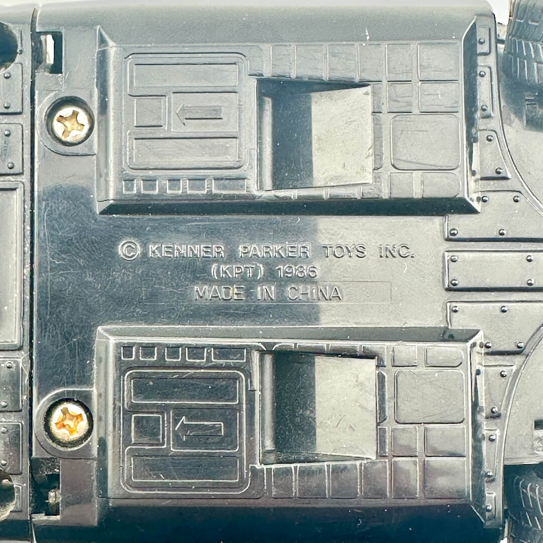 Close-up of the underside of a black plastic toy vehicle, showing molded details, screws, and embossed text.