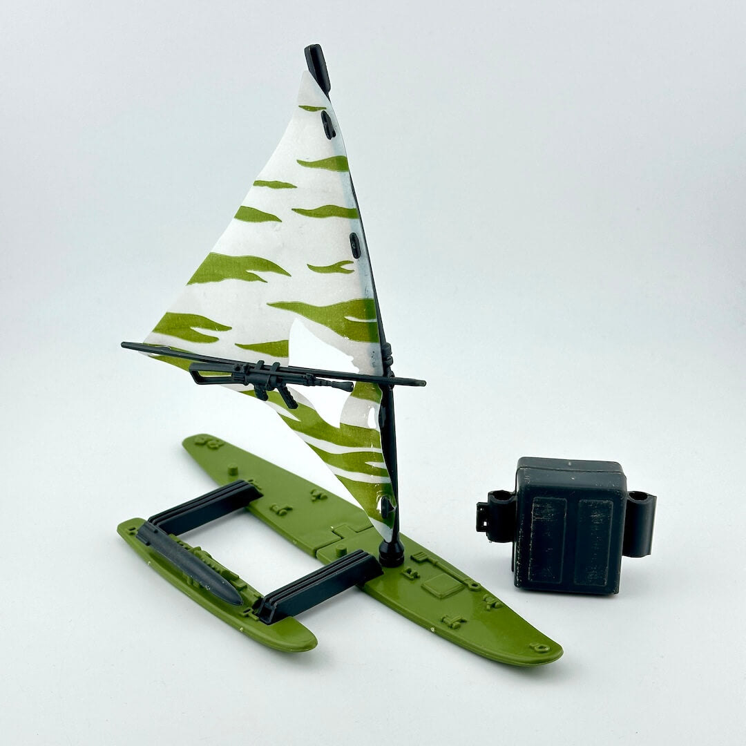 A small green hydrofoil model boat with a white sail featuring olive green camouflage patterns.