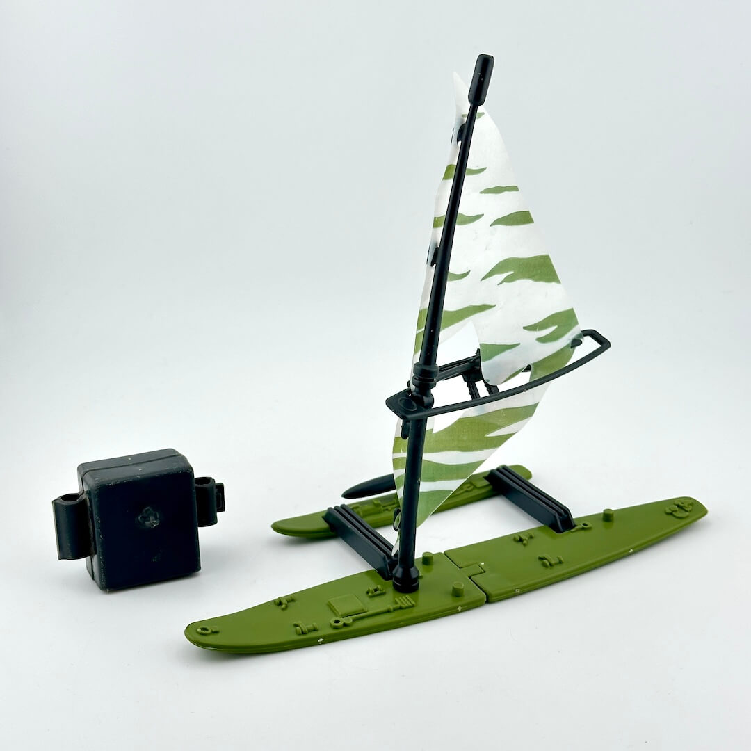 A small plastic toy boat or hydrofoil model with an olive green base, black frame, and a white sail with green camouflage.