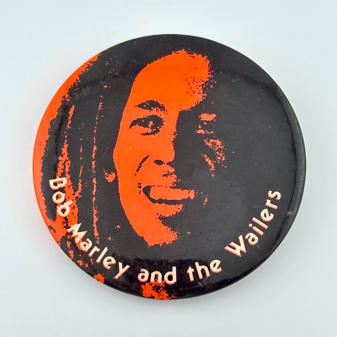 Vintage pin with Bob Marley's face in orange and black and name of his band in white. 