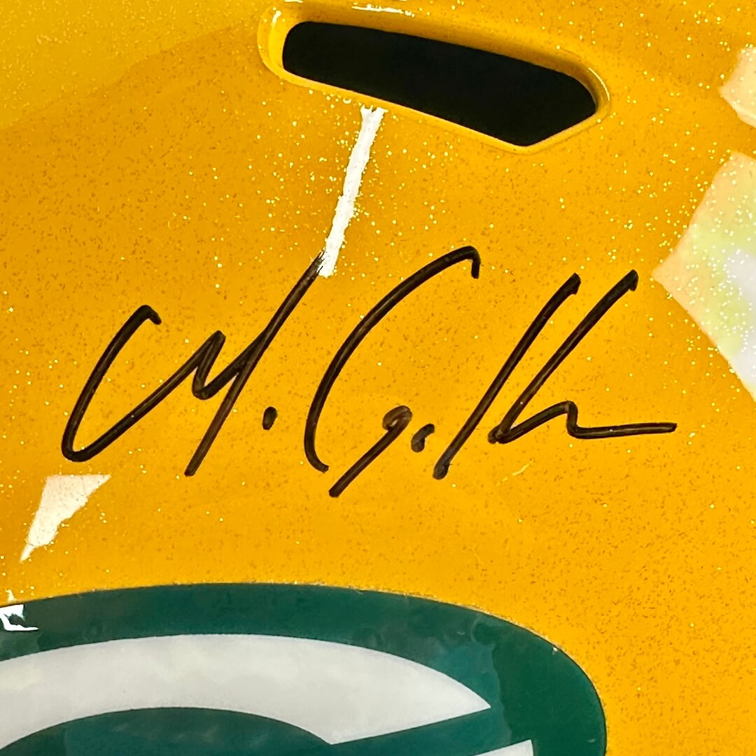 Close-up of a yellow sports helmet with a black autograph across the center and part of a green-and-white logo at the bottom.