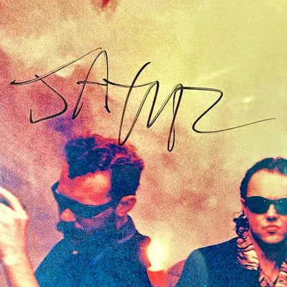 Two people with sunglasses against a colorful background with the signature. 