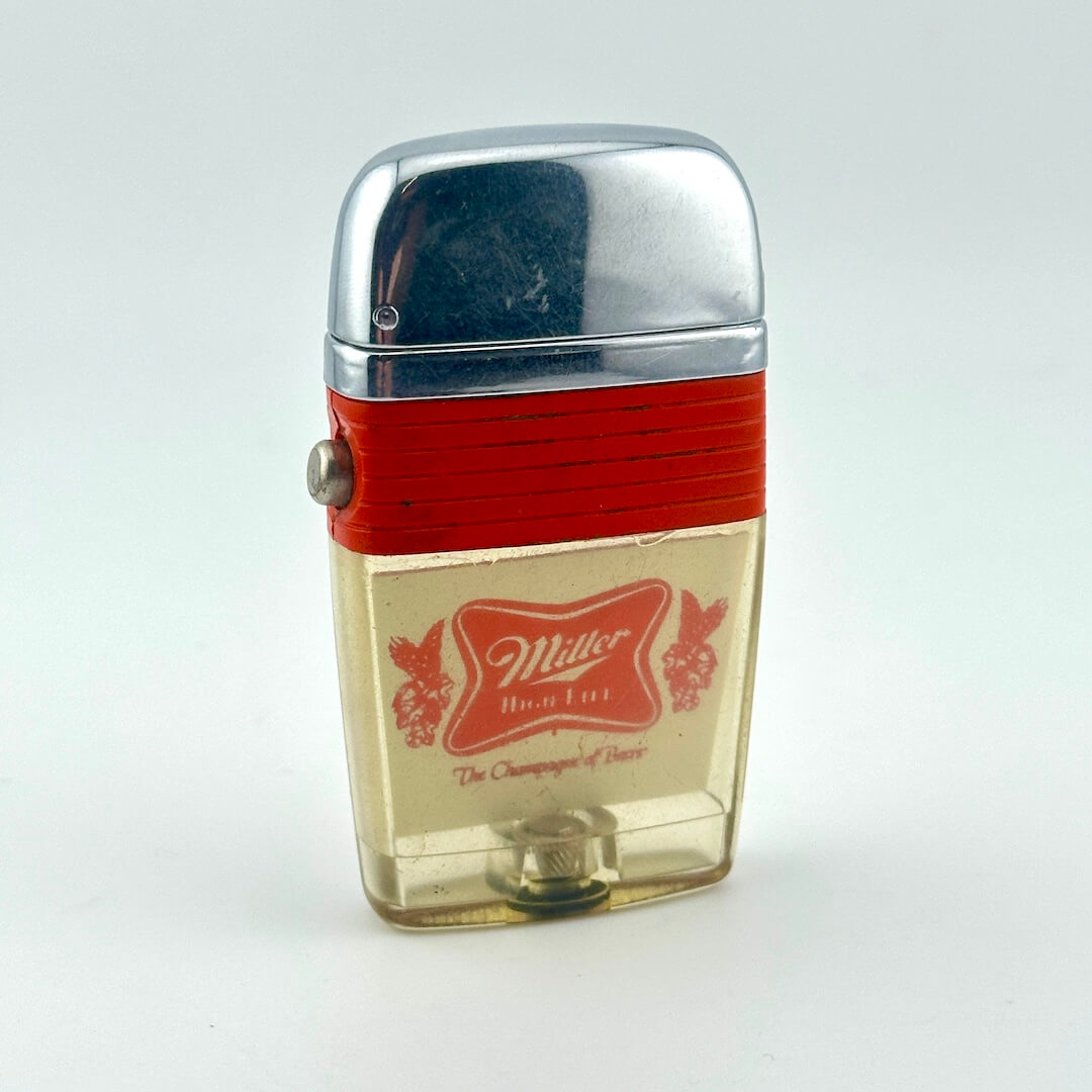 Vintage Miller High Life lighter with a polished chrome cap, a red ribbed band, and a clear lower body showing logo. 