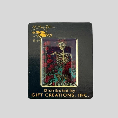 Black card back with hard enamel pin featuring skeleton with crown of roses standing among more roses.
