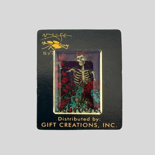 Black card back with hard enamel pin featuring skeleton with crown of roses standing among more roses.