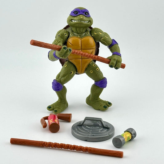 Spotted ninja turtle in purple mask with two bo's, a can of ooze, a trash can lid shield, & fire extinguisher. Front view.
