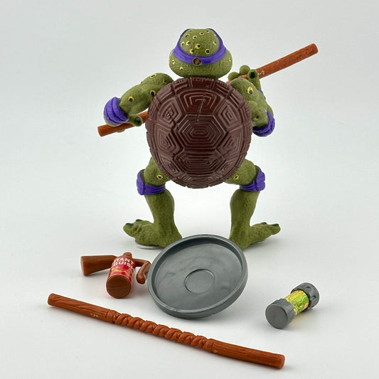Spotted ninja turtle in purple mask with two bo's, a can of ooze, a trash can lid shield, & fire extinguisher. Back view.