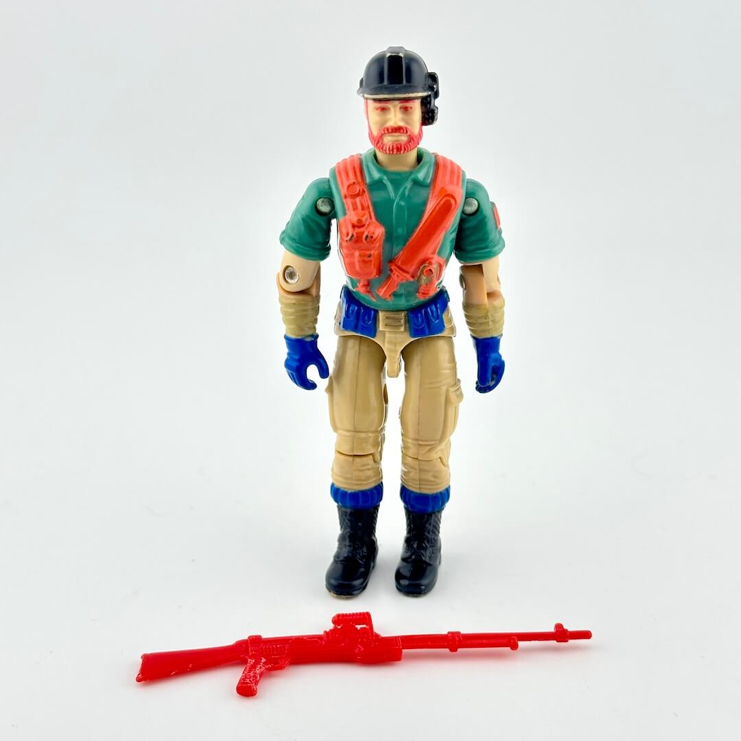 GI Joe action figure with green shirt, black helmet, and brown pants, standing next to red accessory on white background. 