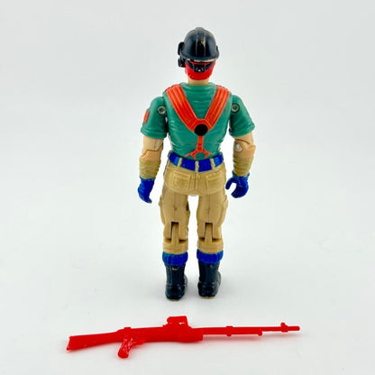 GI Joe action figure with green shirt, black helmet, and brown pants, standing next to red accessory on white background. 