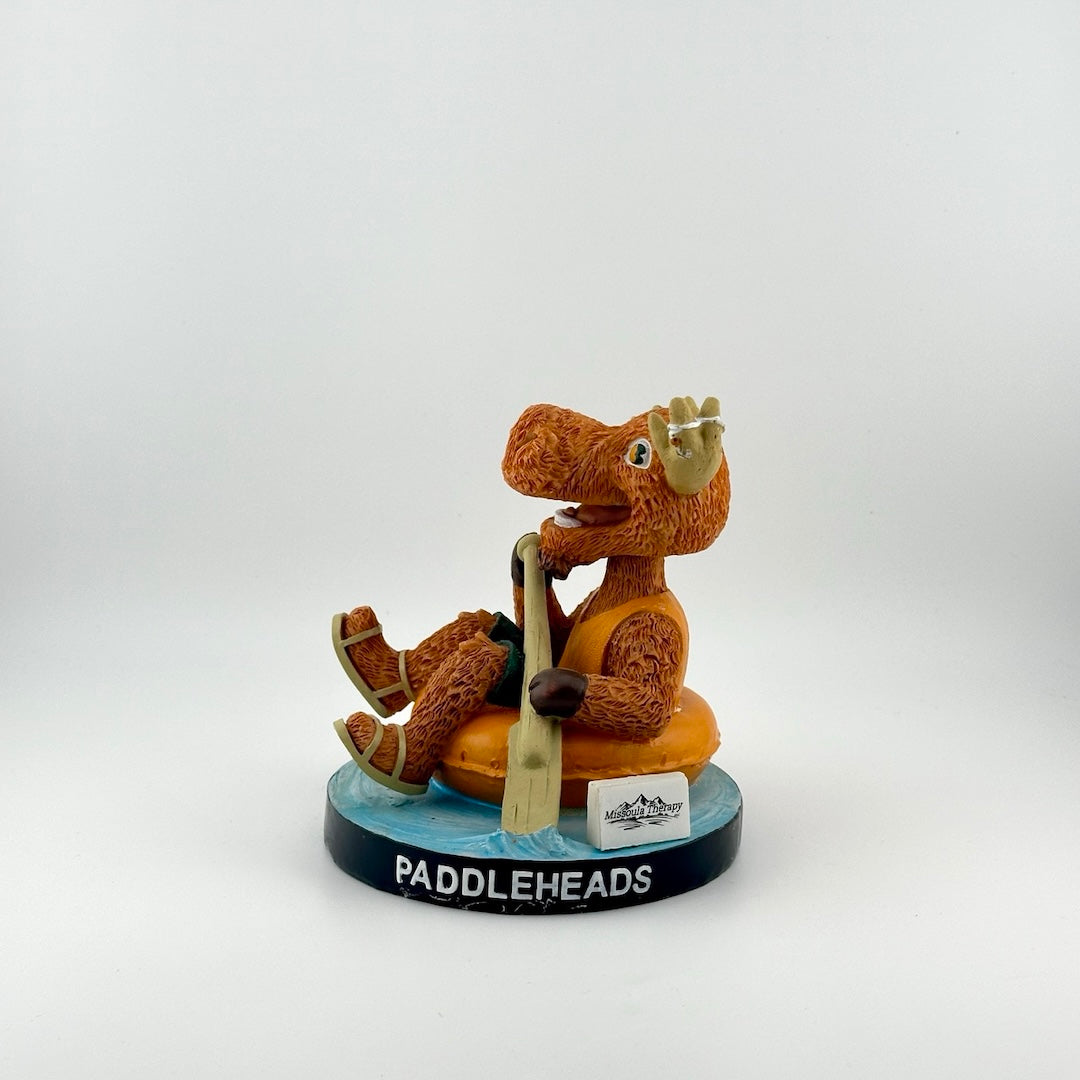 Orange anthropomorphic moose in vest & sandals floating in inner tube with bat as oar. Baseball in horns.