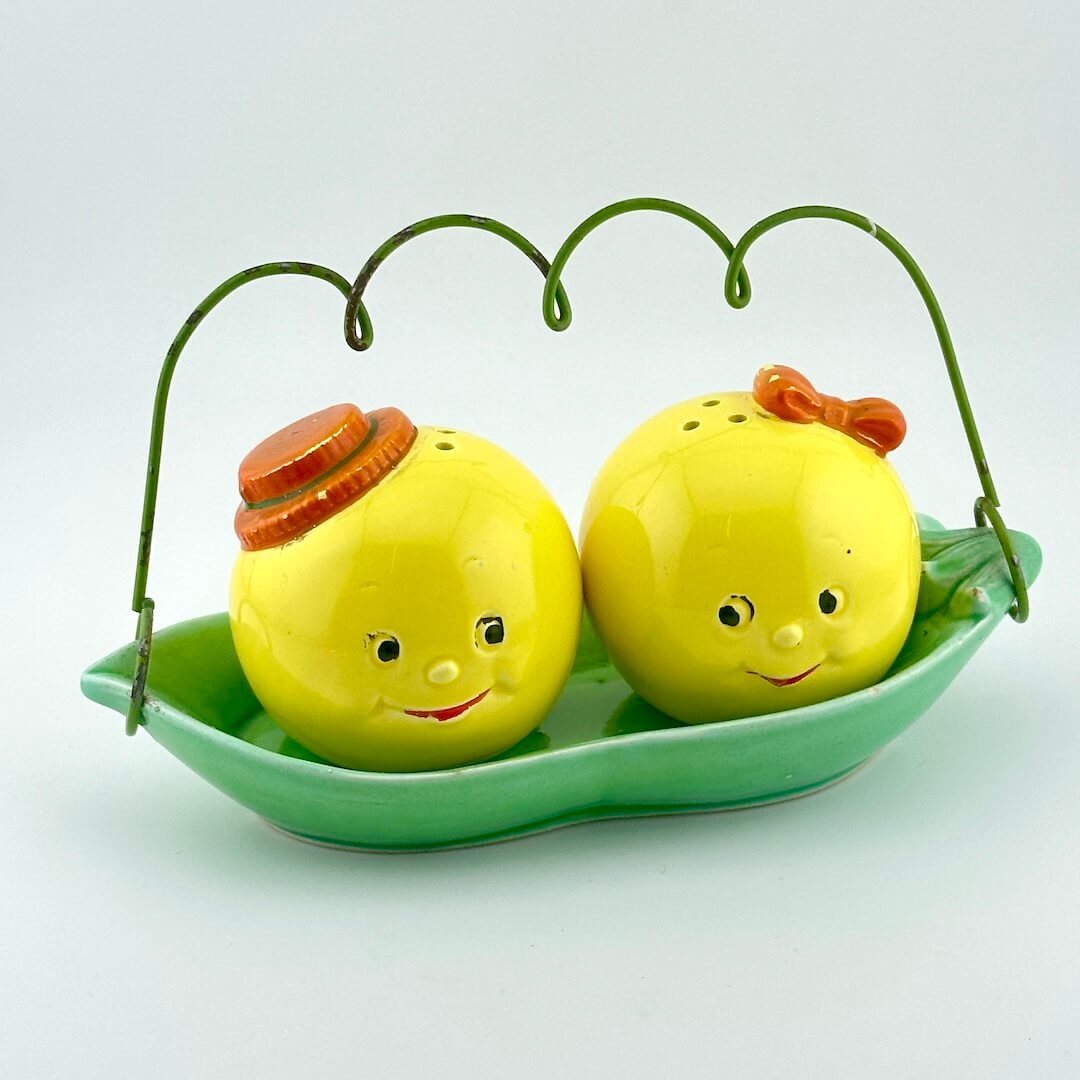 A pair of yellow round salt and pepper shakers with smiling faces, sitting in a green leaf-shaped dish with a green wire handle. 