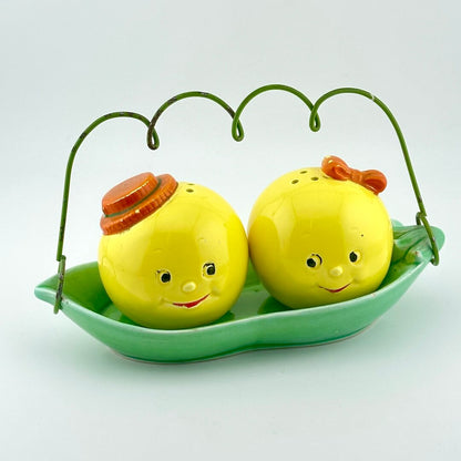 A pair of yellow round salt and pepper shakers with smiling faces, sitting in a green leaf-shaped dish with a green wire handle. 
