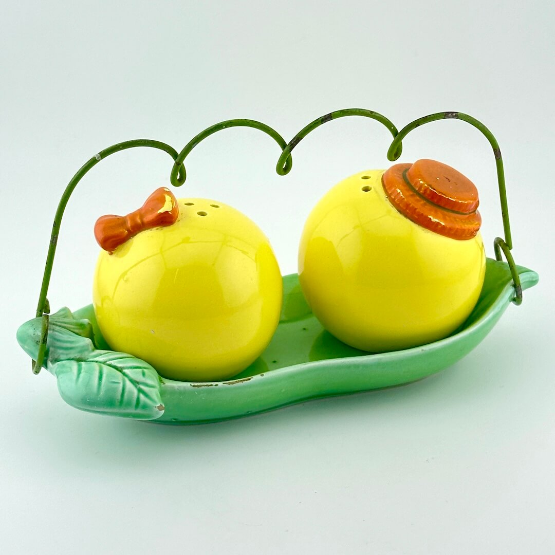 A ceramic salt and pepper shaker set shaped like peas, resting on a green leaf-shaped dish with a wire handle. 