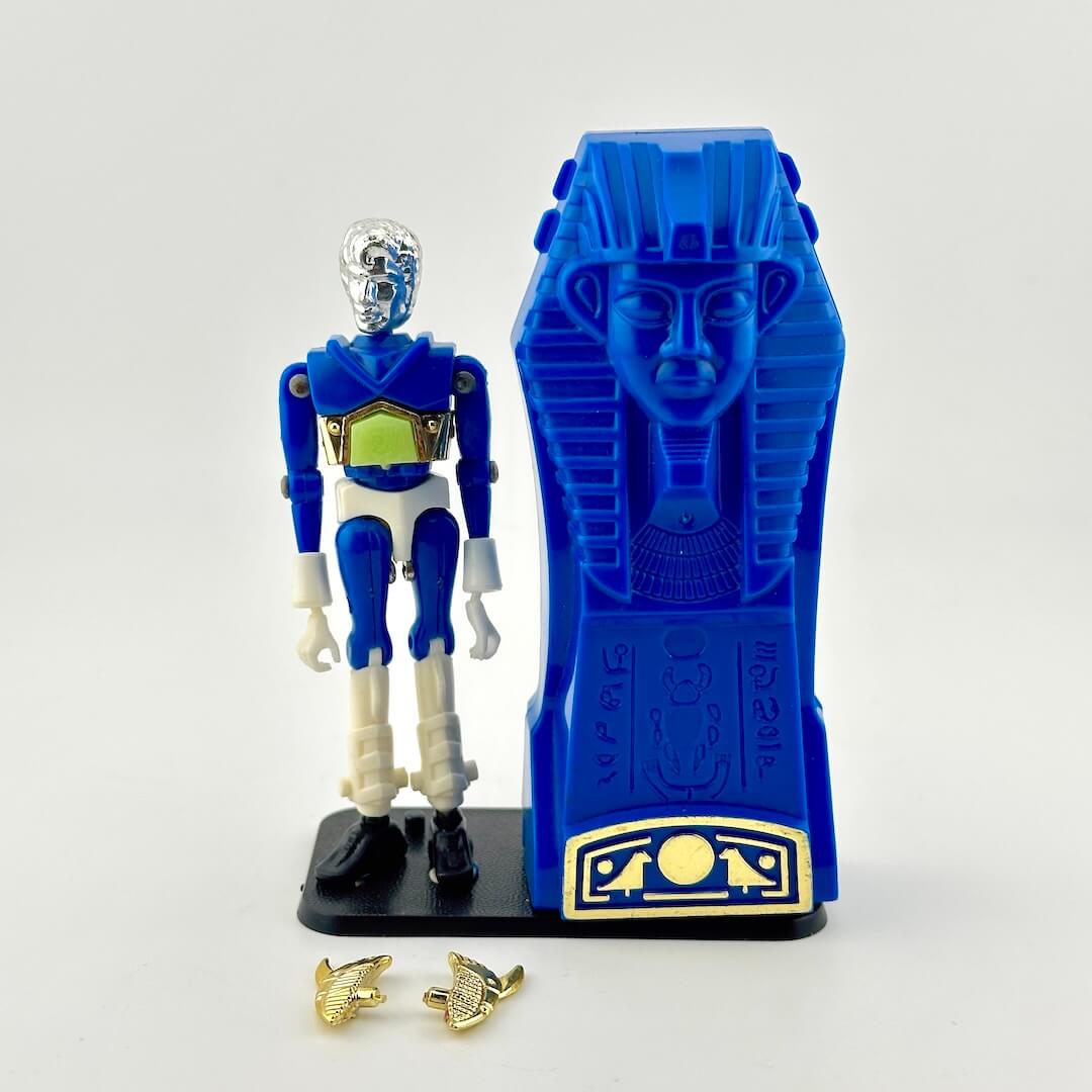 A blue and white action figure standing next to a blue sarcophagus accessory, with two small gold accessories placed in front.