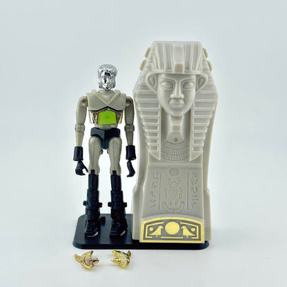 Micronaut action figure standing next to gray sarcophagus with two gold accessories at feet. 