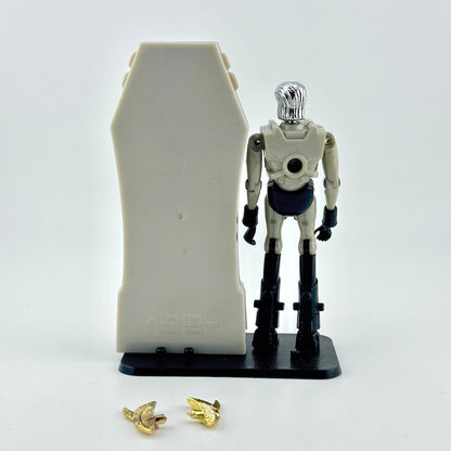 Micronaut action figure standing next to gray sarcophagus with two gold accessories at feet. 