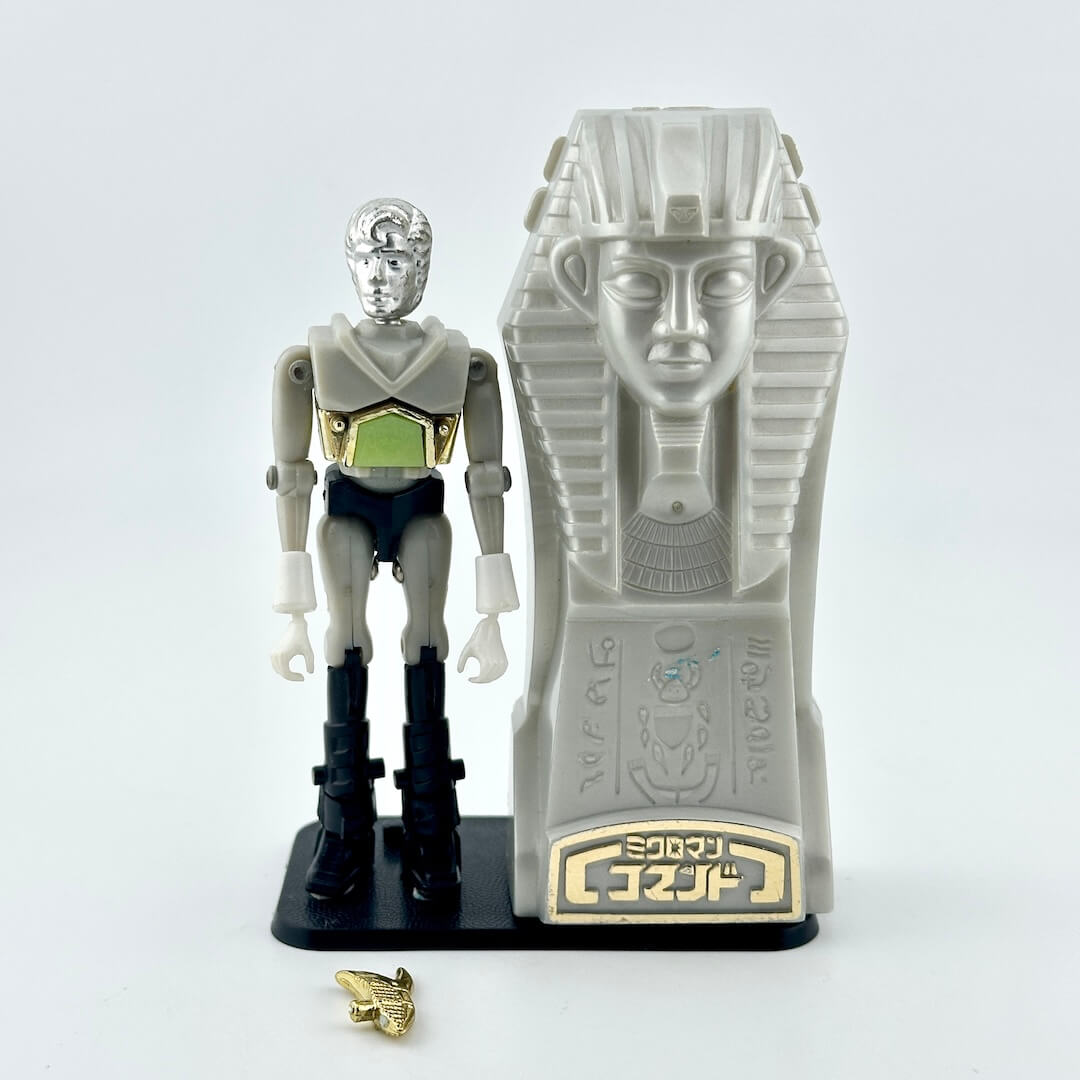 A Micronaut action figure standing next to gray sarcophagus with gold accessories at feet. 