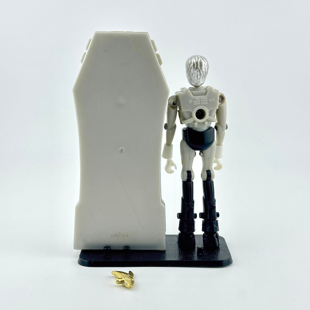 A Micronaut action figure standing next to gray sarcophagus with gold accessories at feet. 