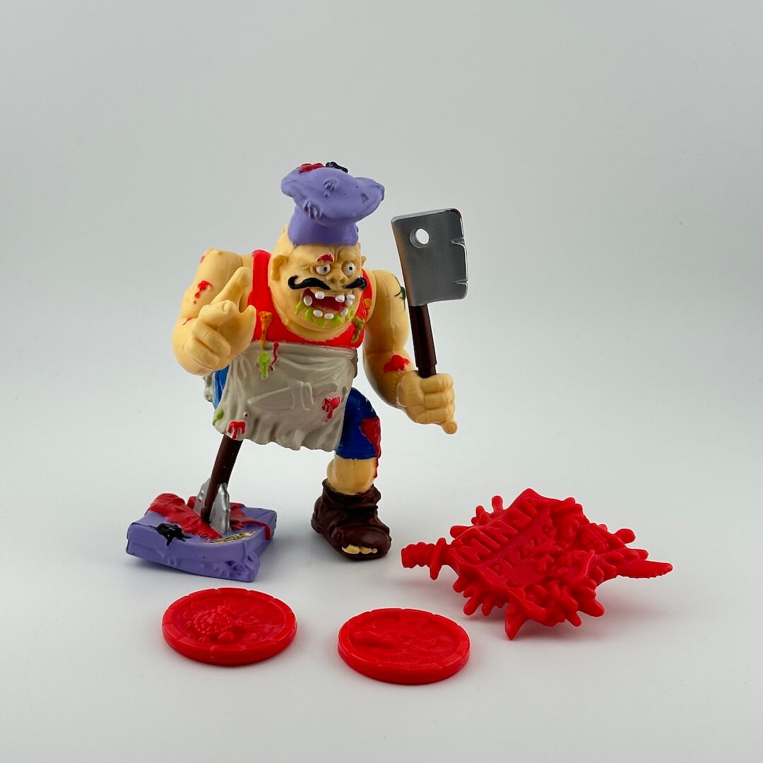 Male figure with peg leg, holding cleaver, in stained apron, blue pants, red tank top & purple chef hat, with red accessories