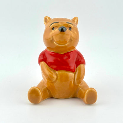 A glossy ceramic figurine of Winnie the Pooh sitting down, wearing his signature red shirt, on a clean white background.