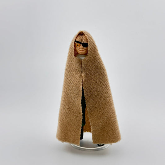 Alien figure wearing tan felt cape, with a peach flesh tone, orange eye with slit pupil & black eye-patch on other side.