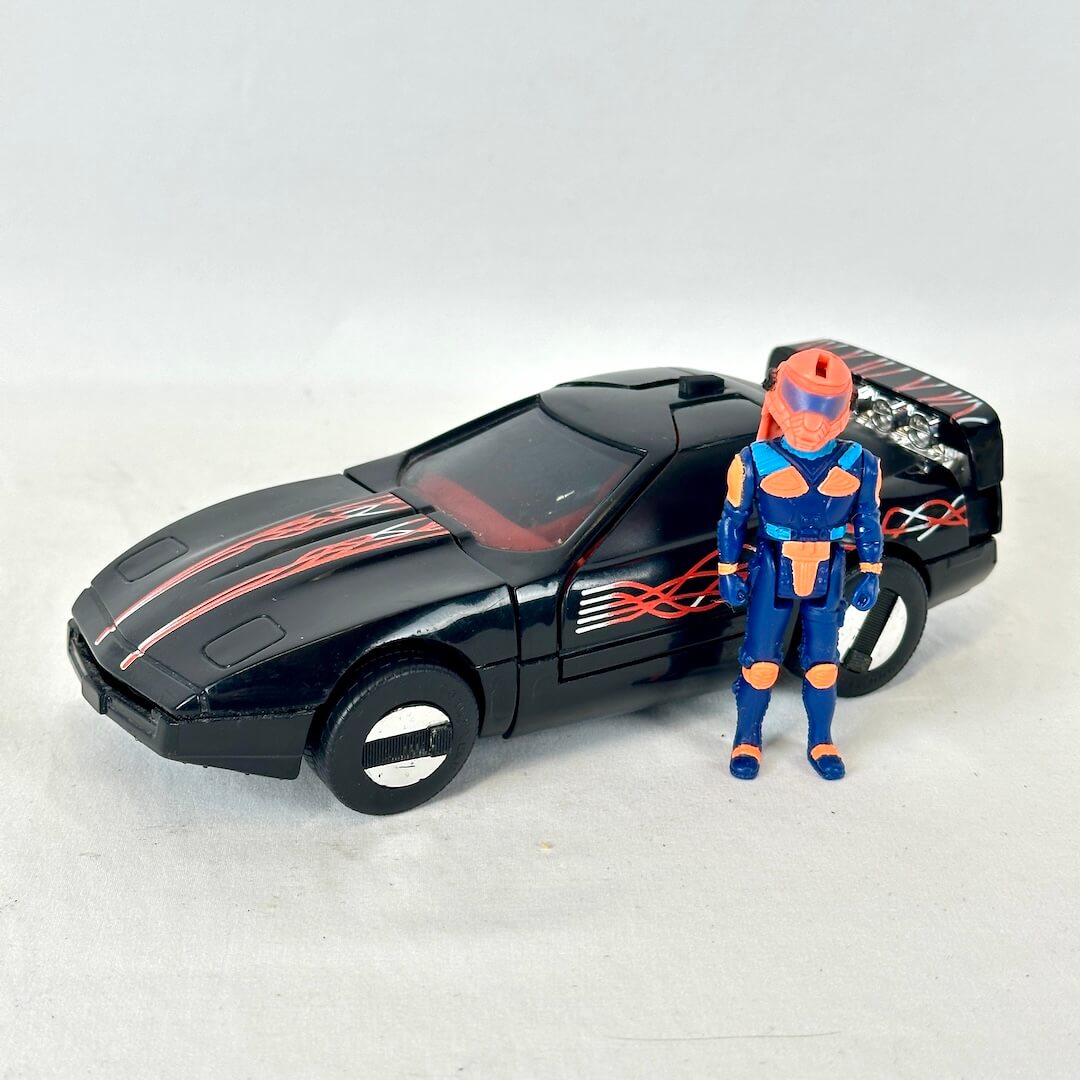A black toy sports car with red flame decals, shown next to a blue and orange action figure on a light gray background.
