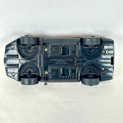 An upside-down black toy car chassis with four black wheels and small gold-colored screws. 
