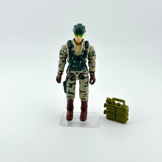 Action figure in camouflage uniform with helmet and boots, standing next to a small green accessory on a white background