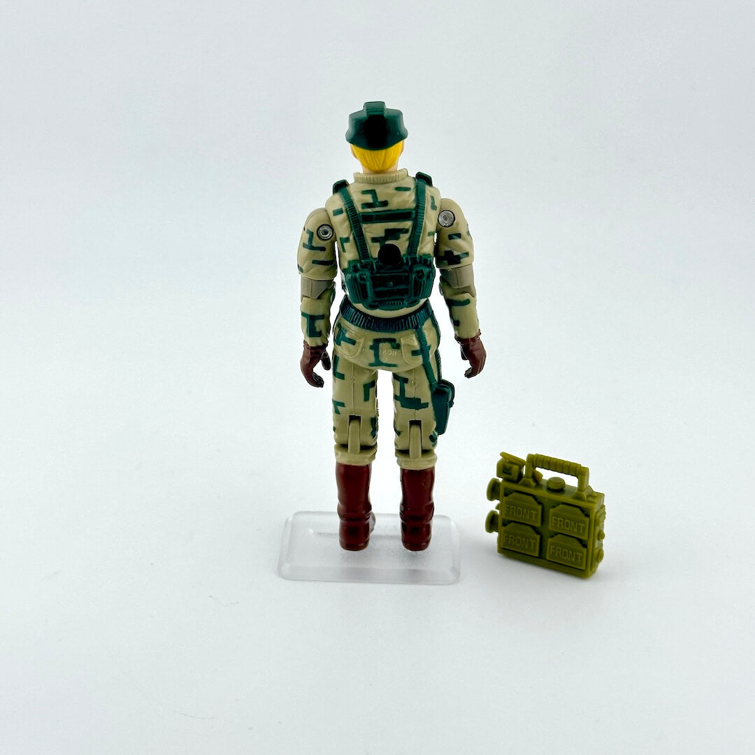 Action figure in camouflage uniform with helmet and boots, standing next to a small green accessory on a white background